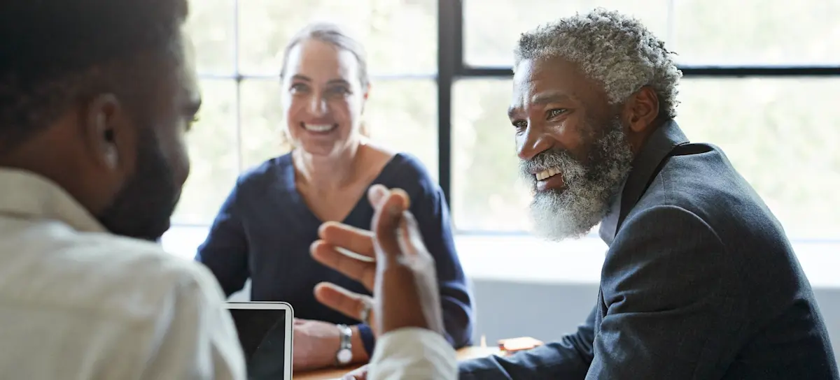 [Featured image] A person smiles at their colleague while listening to them speak. In the background, another person sits at the table listening and smiling at the story. All three colleagues are using their soft skills to communicate, listen, engage, and relate to one another.
