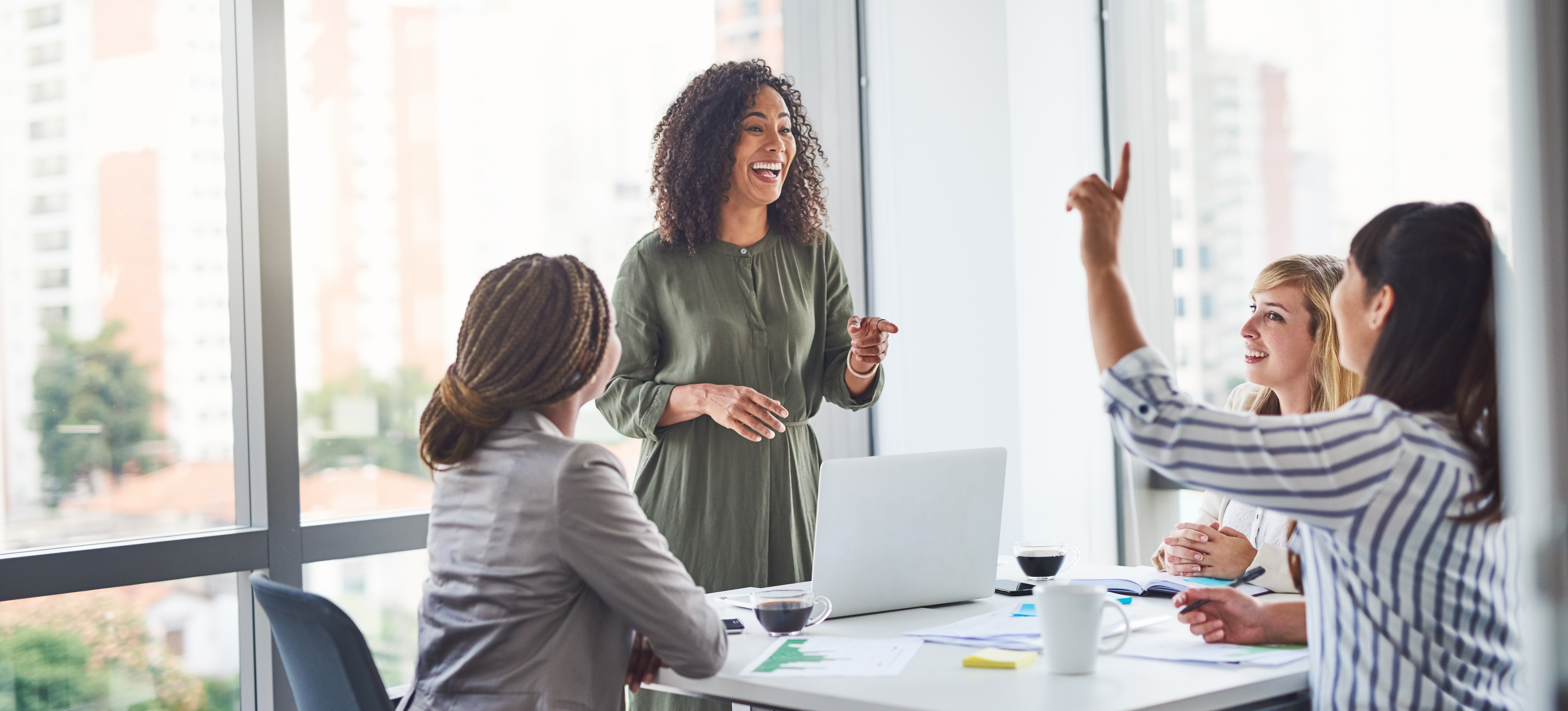 [Featured Image] Working together, a smiling team of four women use their problem-solving skills to overcome a business hurdle, as they gather around a desk in an office.
