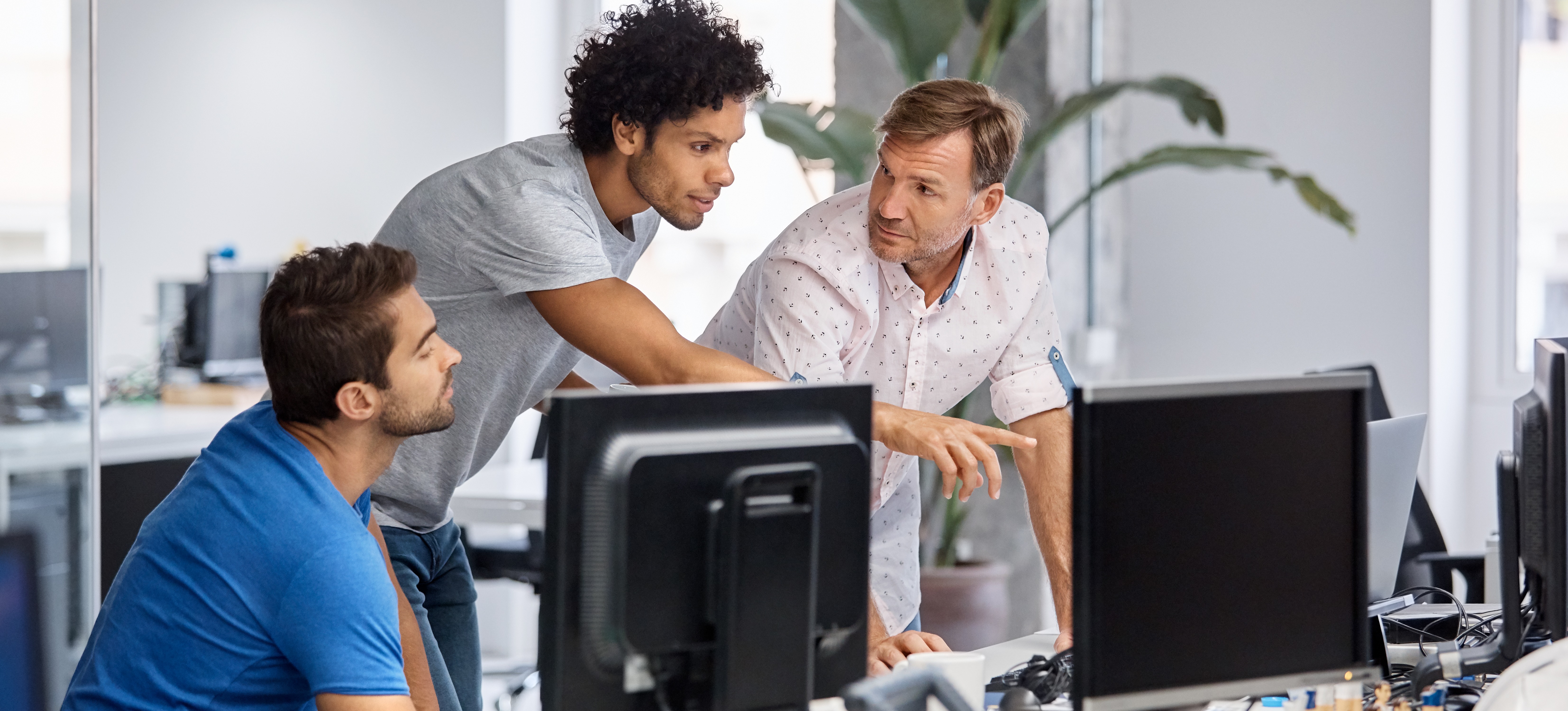 [Featured Image] Three programmers discuss cloud infrastructure in an office setting while one points to a computer monitor.