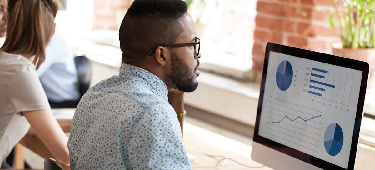 [Featured image] A digital marketing expert wearing glasses sits at a shared desk and reviews the performance of marketing channels on his desktop monitor.