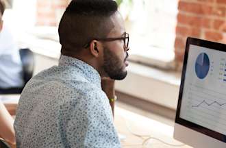 [Featured image] A digital marketer in glasses sits at a shared desk and reviews marketing channel performance on their desktop monitor. 