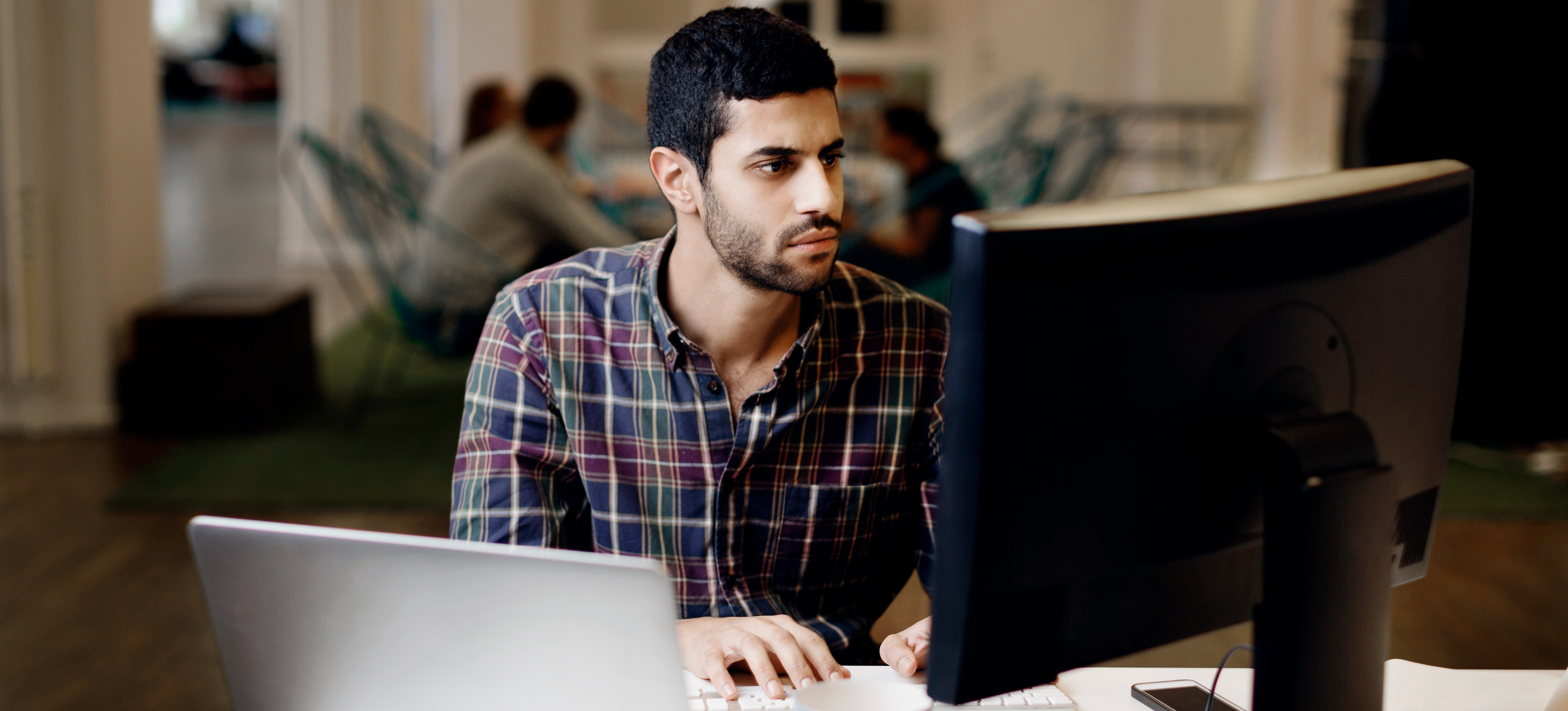 [Featured Image] A data analyst works with data types in Java on their computer while colleagues have a meeting around a table in the background.