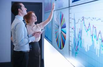 [Featured Image] Two dala analysts examine charts and graphs projected on a white smart board.