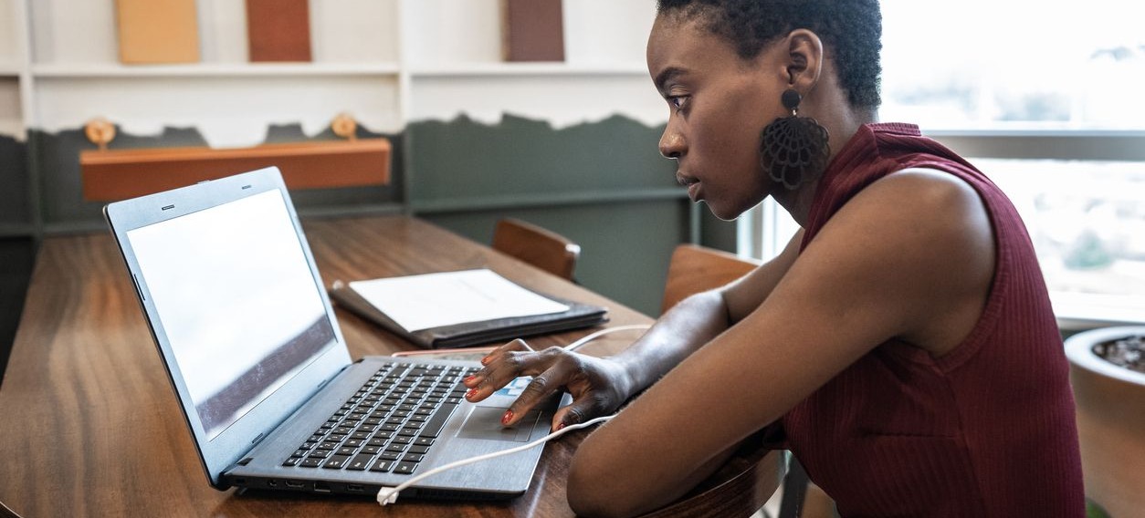 [Featured Image] A woman sitting at a desk using her laptop with a tablet placed on the table. 