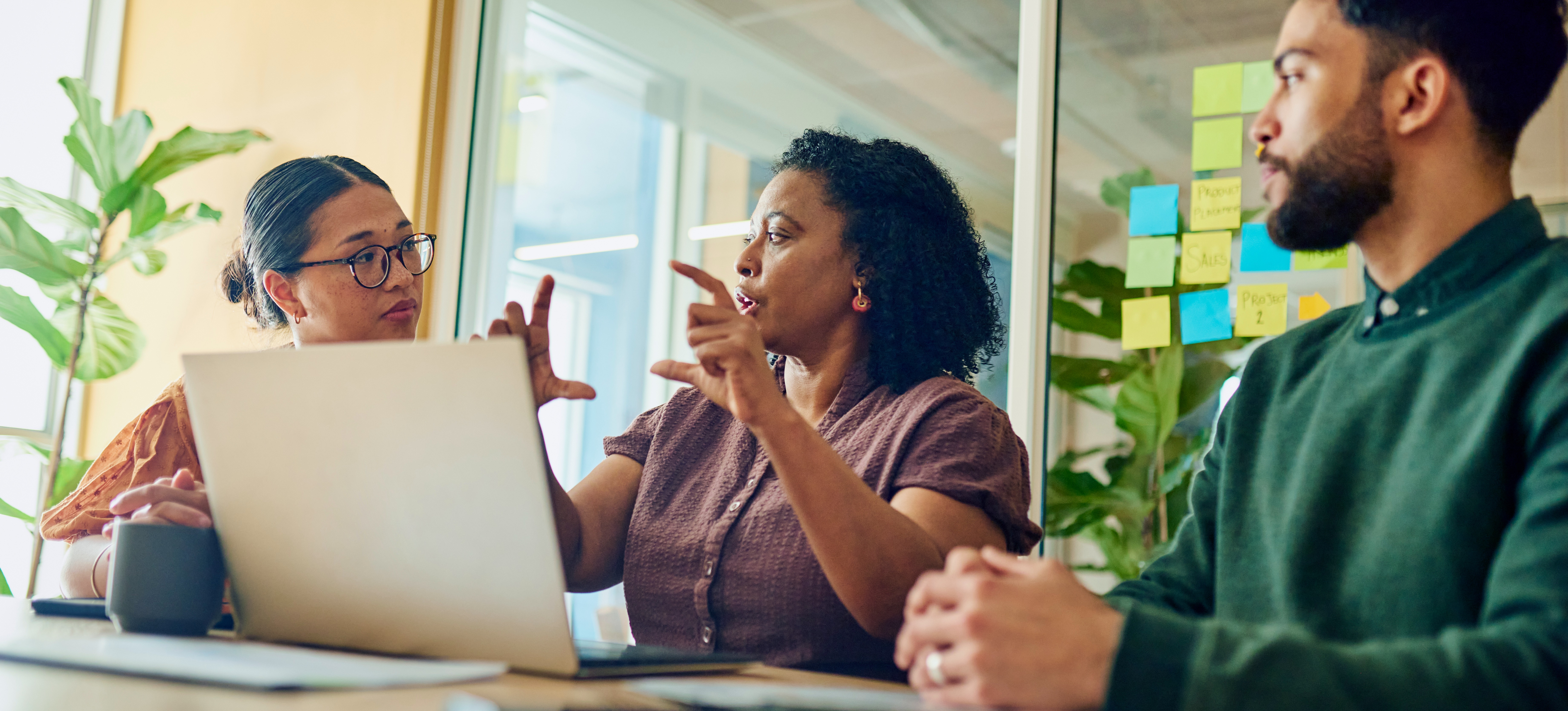 [Feature Image] Three marketing professionals collaborate around a laptop computer on a table in a conference room by brainstorming their strategy for a visual campaign.
