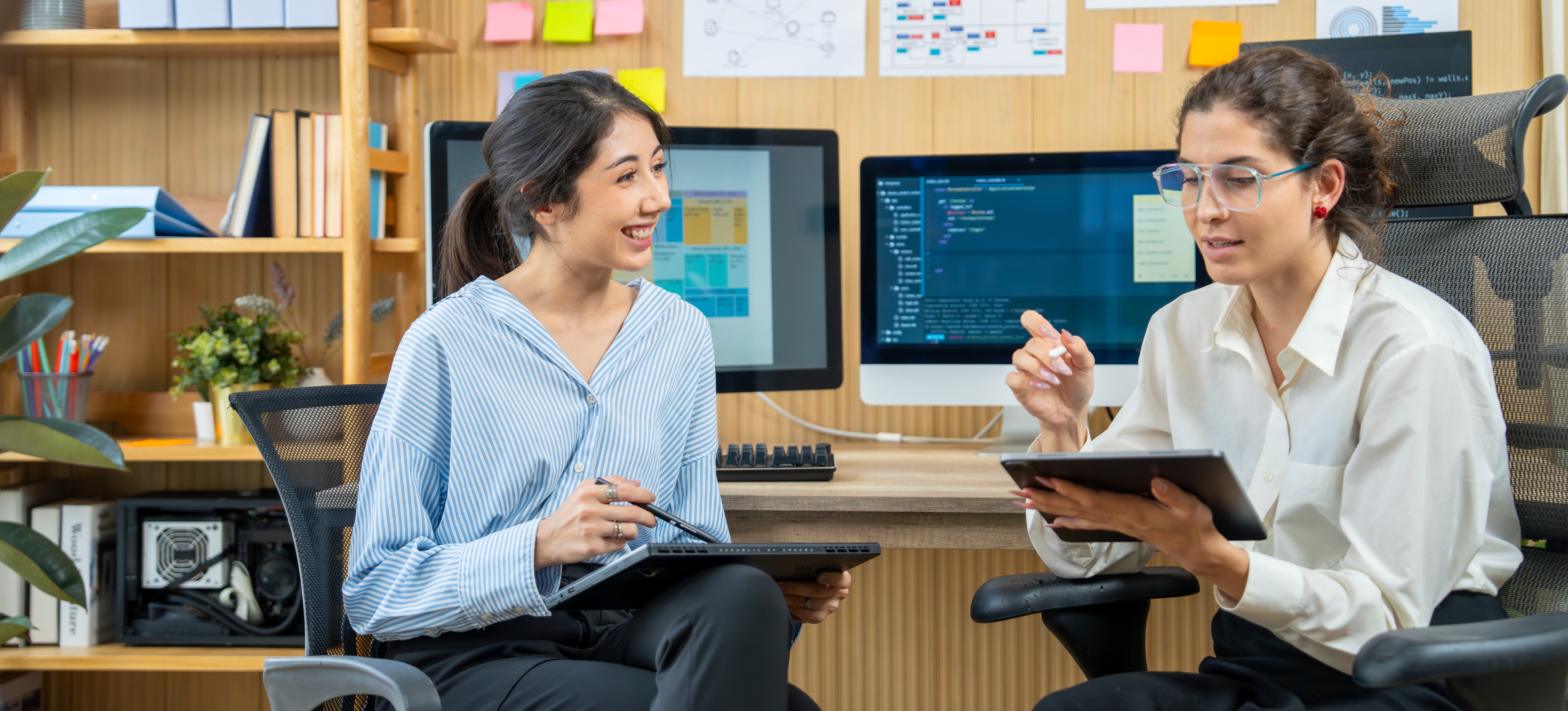 [Featured text] Two information technology specialists leverage AI in networking to monitor performance and analyze data. They are sitting with their backs to two computer monitors filled with charts and screens that showcase productivity and other data.
