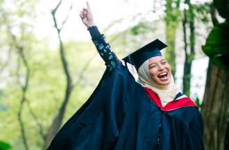 [Featured image] A young woman in a hijab and graduation gown raises her finger excitedly in the air. 