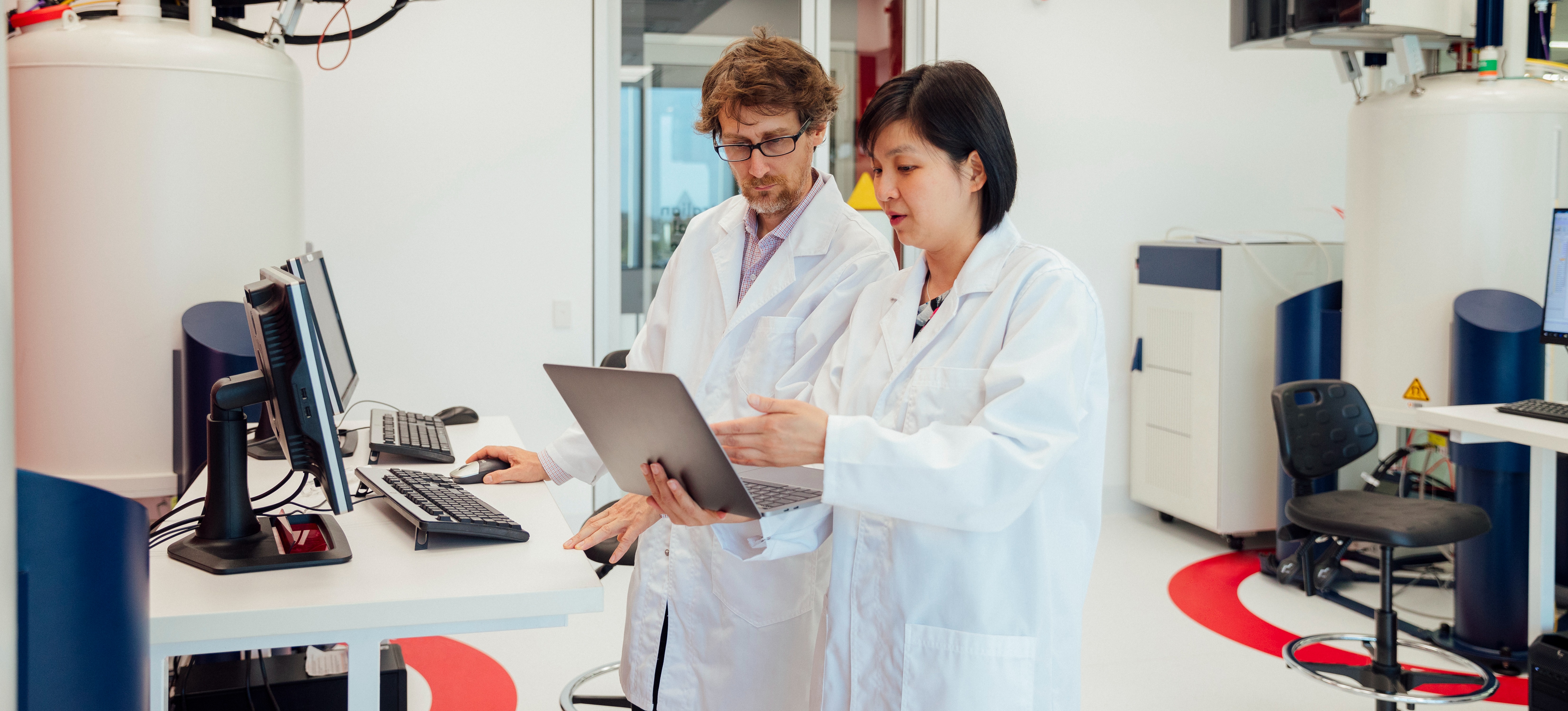 [Featured image] Two health information managers stand in a white room, looking at patient data on a laptop computer while standing in front of two desktop computers
