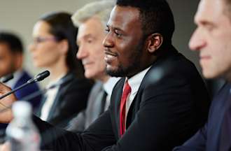 [Featured image] A group of professionals in business suits sit in front of microphones at an international press conference.