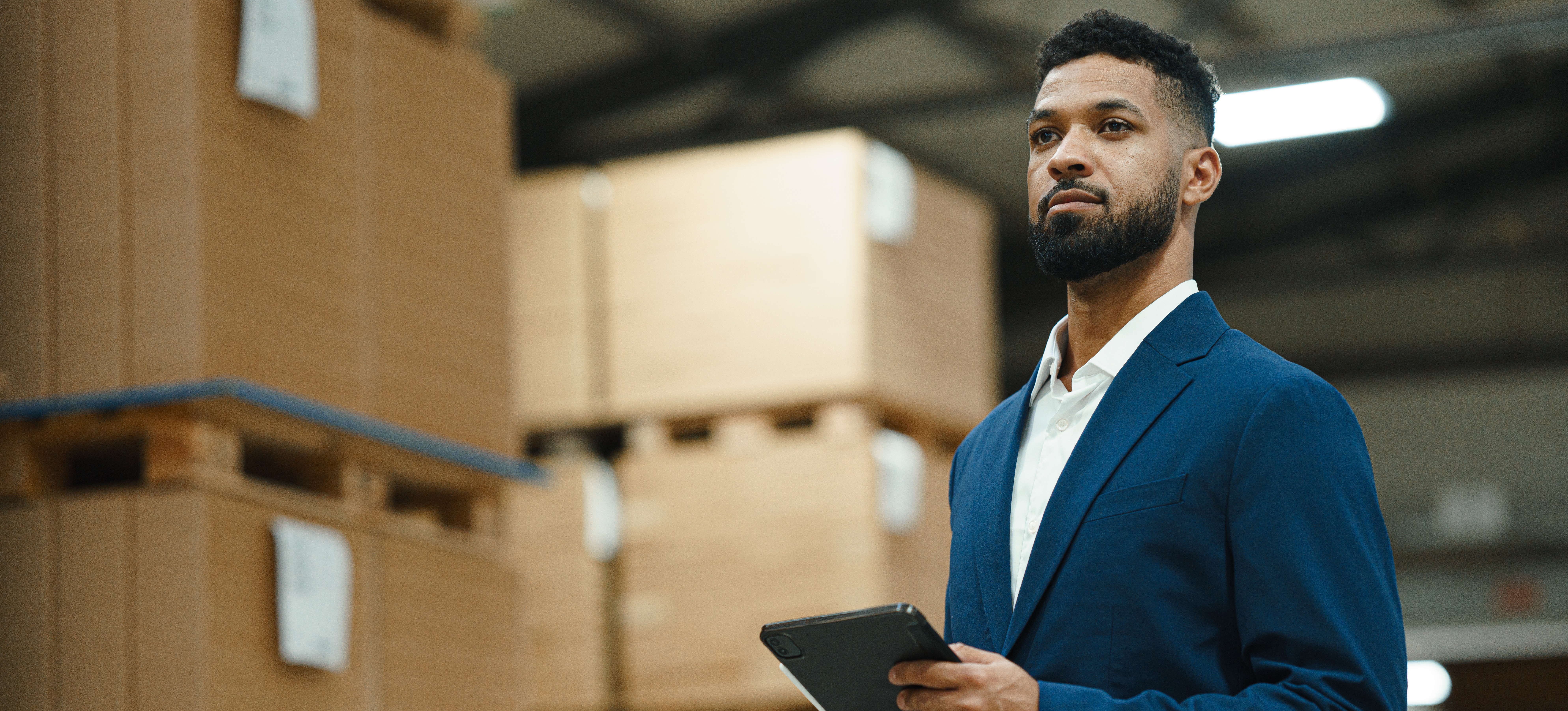 [Featured Image] A product manager holding a tablet stands in a warehouse full of boxed inventory.
