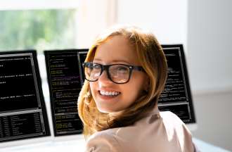 [Featured Image]:  A coder, sitting at her desk, working with two desktop computers, is preparing to begin a coding bootcamp.