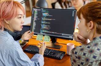 [Featured Image] A young web developer sitting in front of a computer and discussing their coding with a coworker.