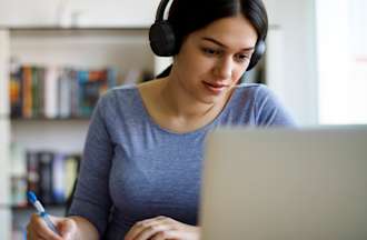 [Featured image] A young person wears over-the-ear headphones while working with a pen and paper in front of their laptop. 