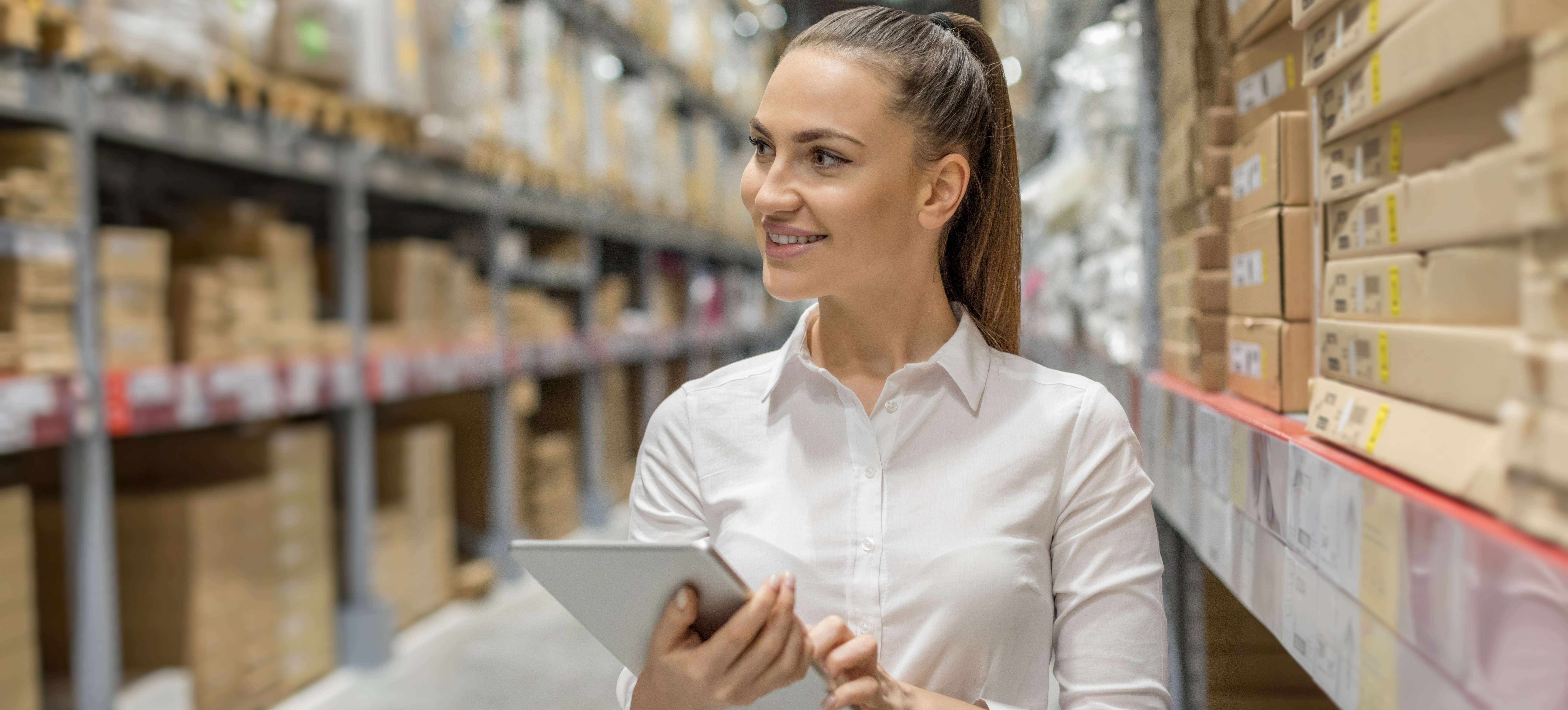 [Featured Image] An operations manager walks through a warehouse full of boxes holding a tablet to check inventory levels. 
