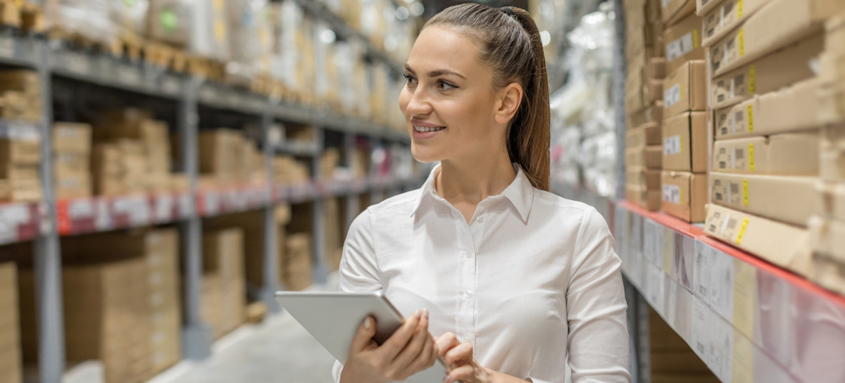 [Featured Image] An operations manager walks through a warehouse full of boxes holding a tablet to check inventory levels. 

