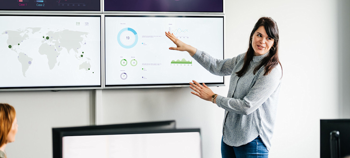 [Featured Image] A business person is leading a meeting displaying four screens, each displaying visualizations created with AI data analysis tools. 
