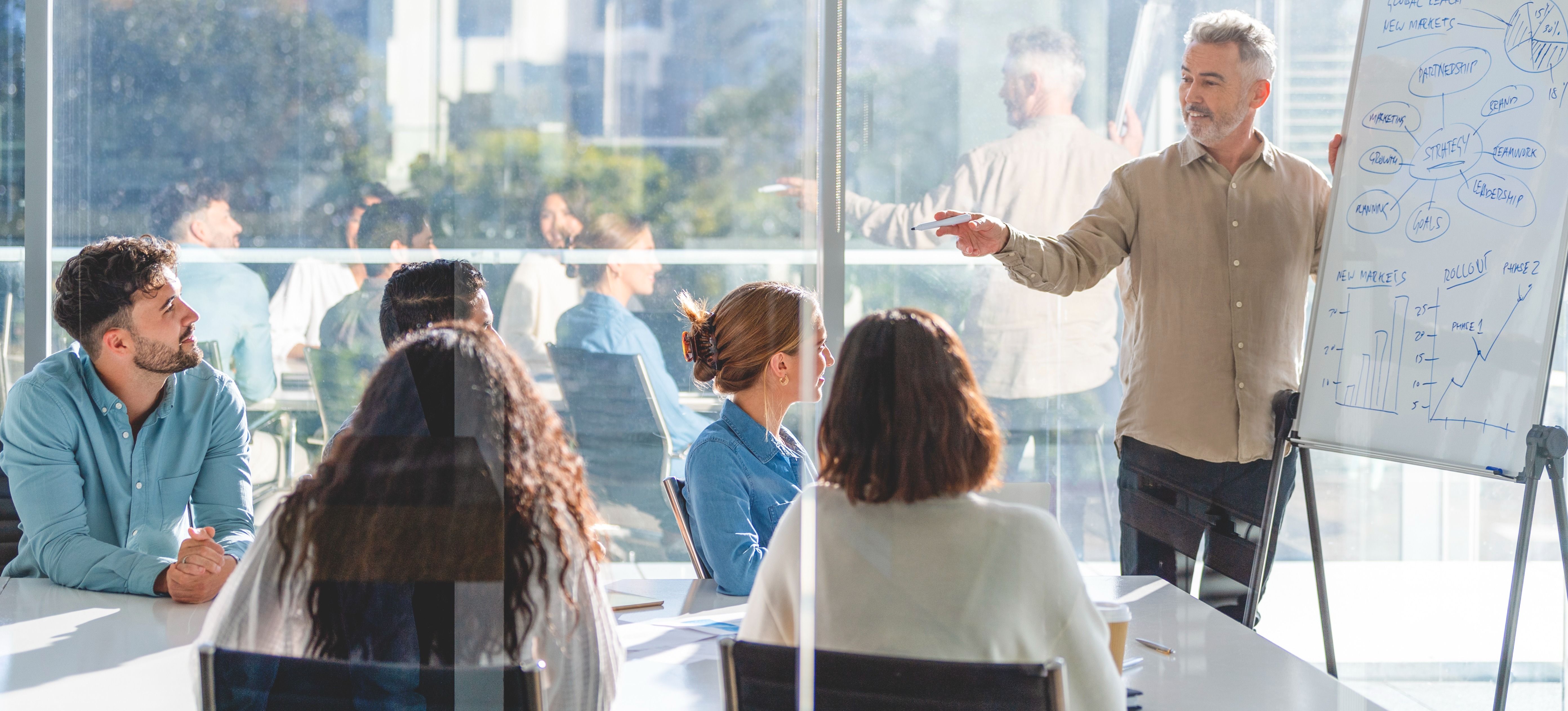 [Hauptbild] Ein Trainer steht vor einer Gruppe von Führungskräften und hält in einem sonnigen Konferenzraum eine Schulung zum Thema Managemententwicklung, wobei er eine weiße Tafel verwendet.