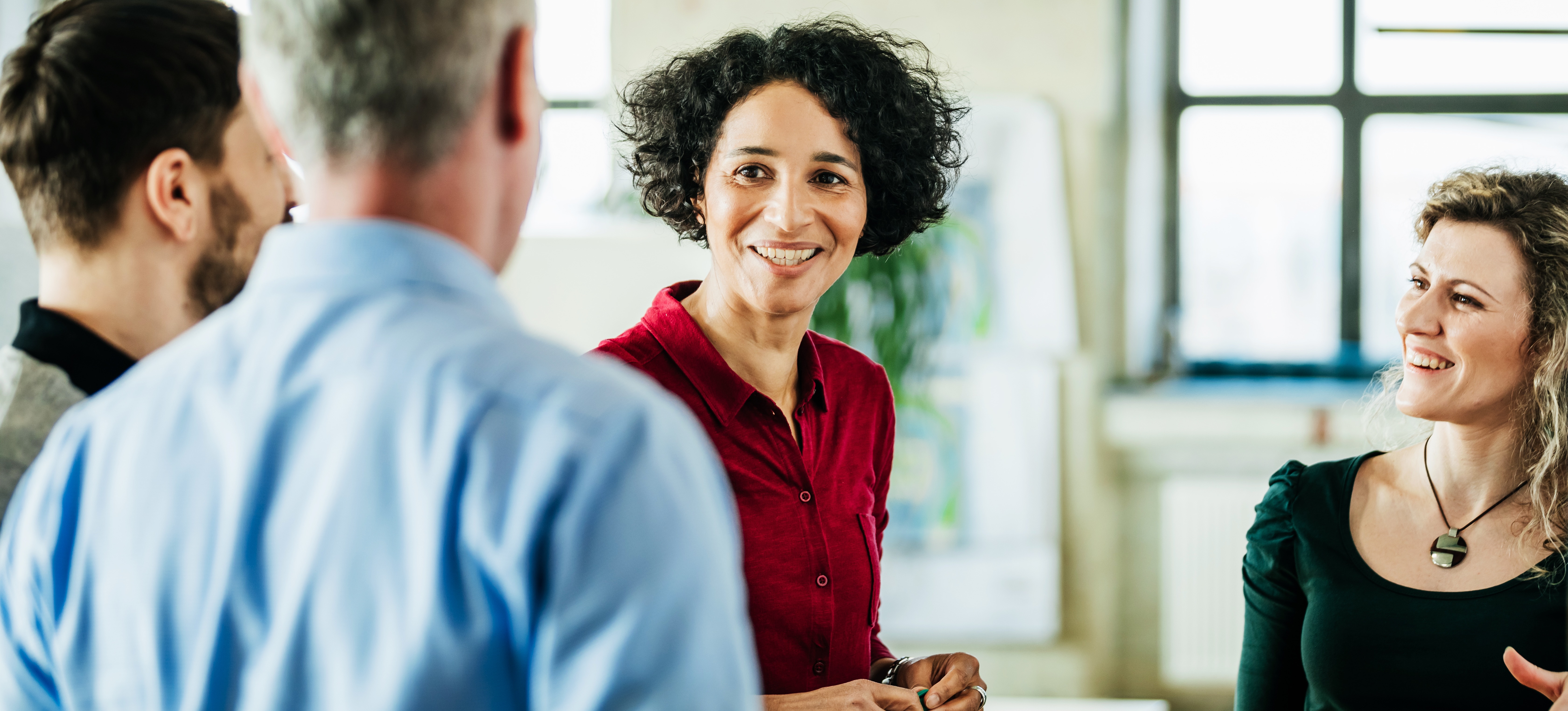 [Featured Image] A smiling business process analyst speaks with her happy colleagues while standing in a sunlit, spacious board room. 
