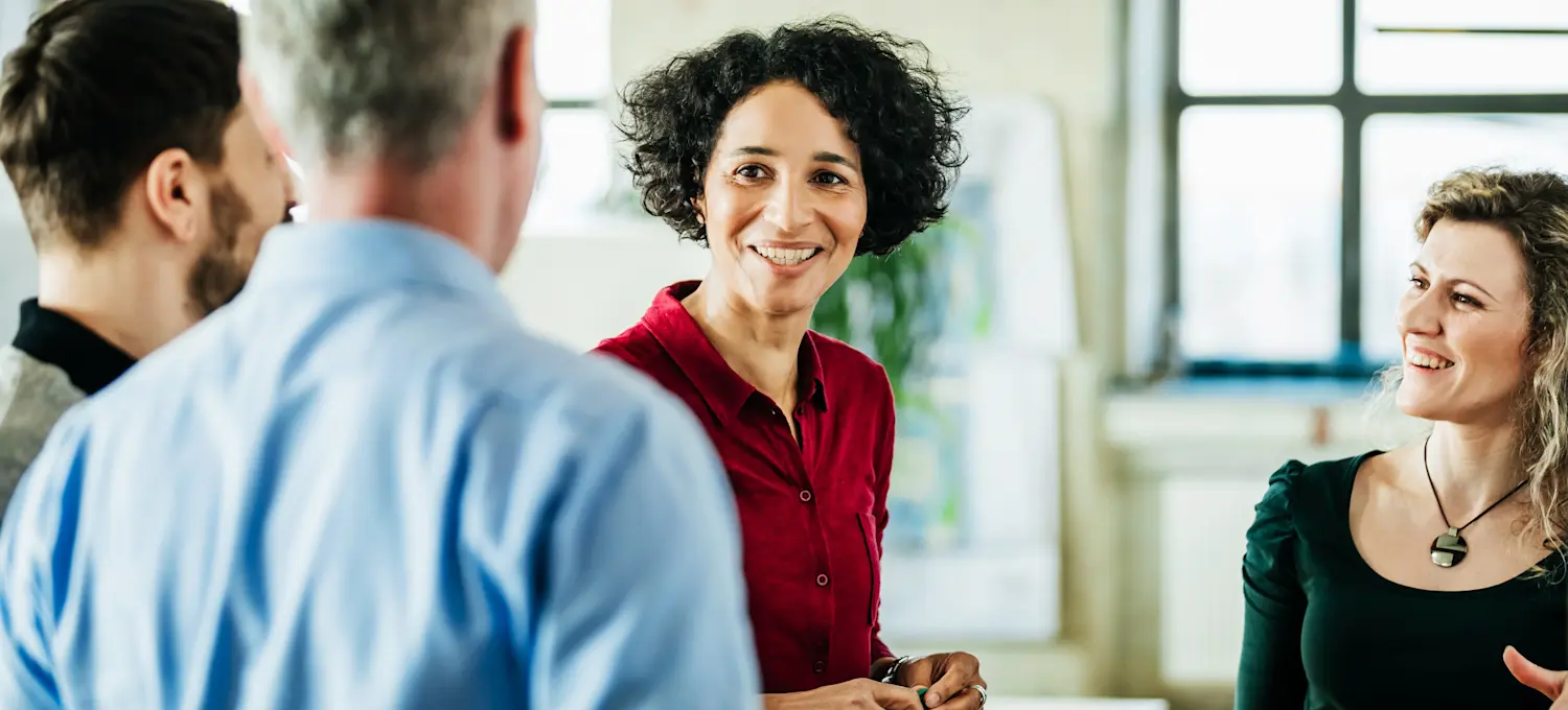 [Featured Image] A smiling business process analyst speaks with their happy colleagues while standing in a sunlit, spacious board room.