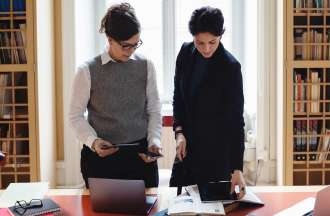 [Featured image] Two accountants examine an accrued expenses journal at a table in a library.