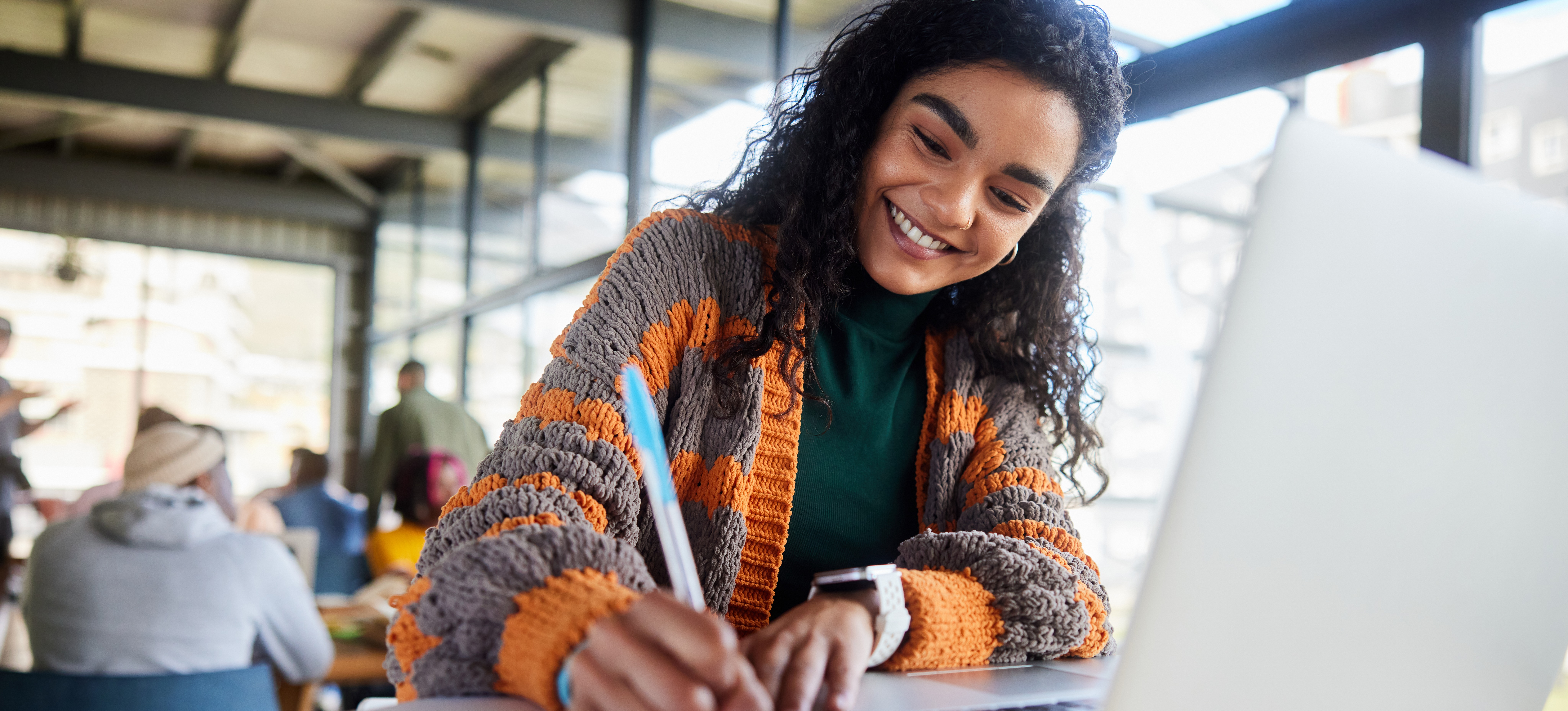 [Feature Image] A dark-haired learner smiles as they complete coursework that’s part of their general studies degree program while working on a laptop in a windowed room with other general studies learners.
