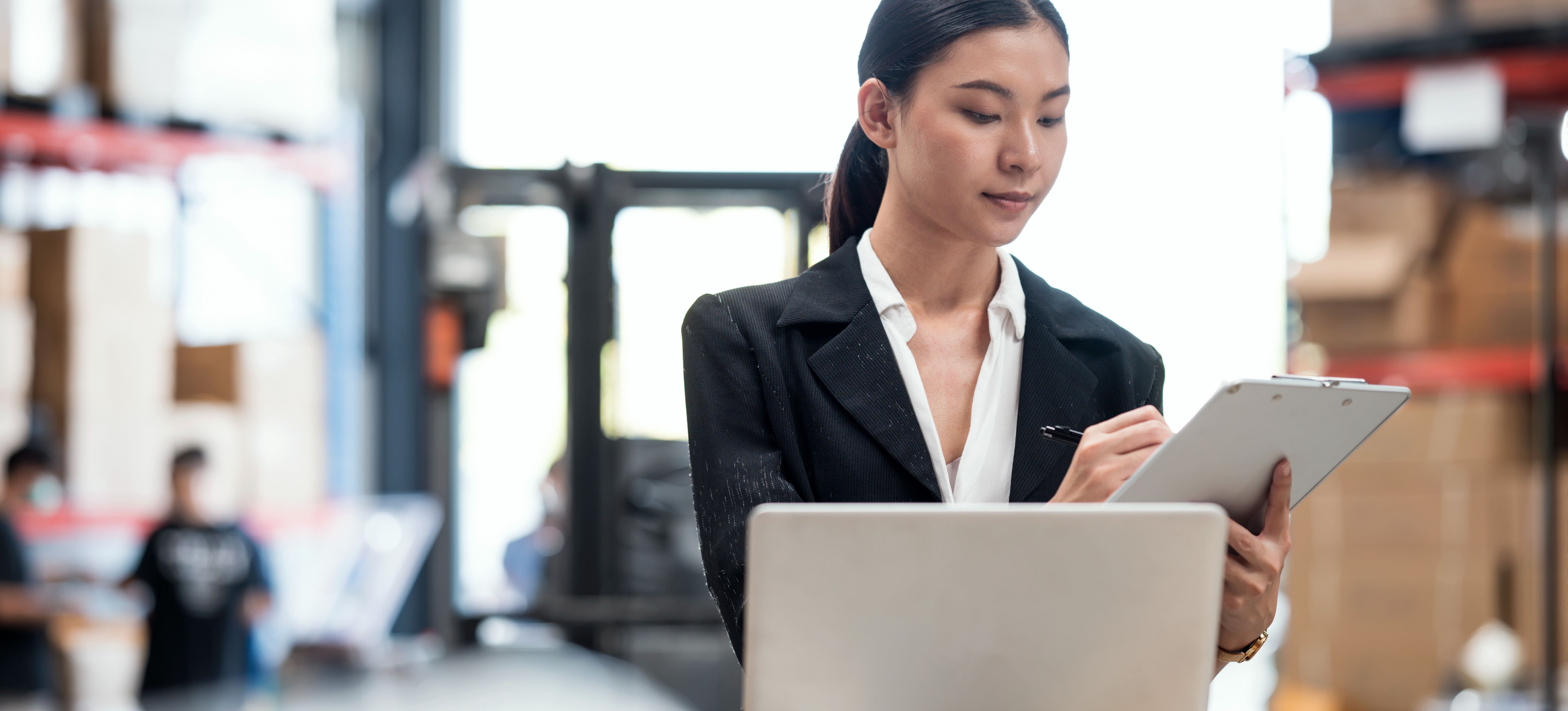 [Featured Image] A worker in a warehouse uses a computer and clipboard as they start a new job in cyber security in the retail industry.
