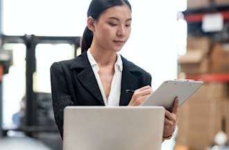 [Featured Image] A woman in a warehouse uses a computer and clipboard as she starts a new job in cyber security in the retail industry.
