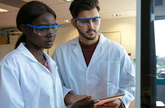 [Featured Image] Two health care workers wear protective glasses and lab coats and stand in front of a medical instrument.