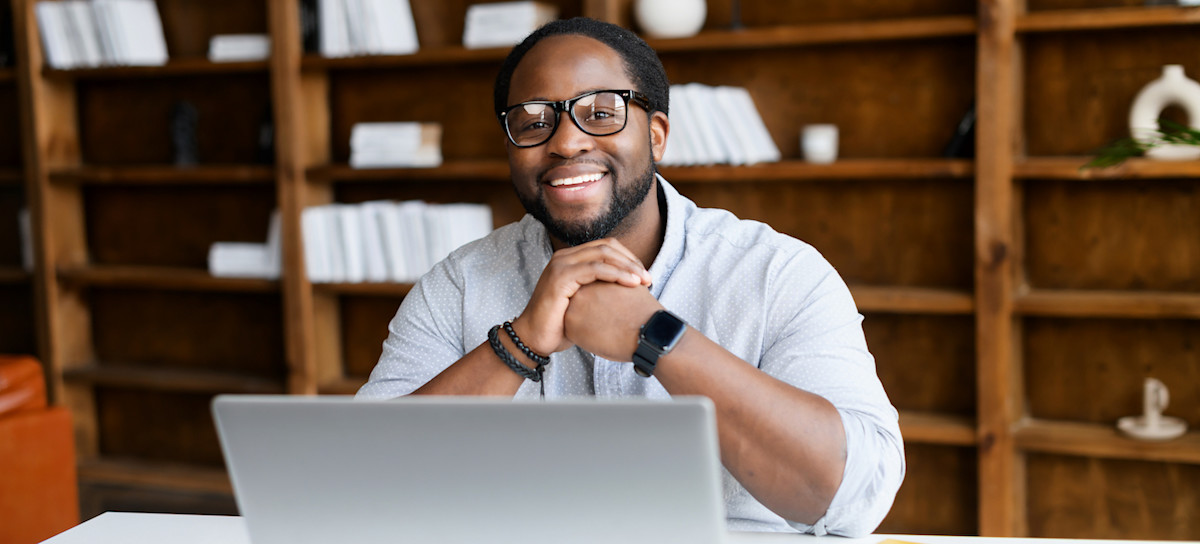 [Featured image] A person in classes, sitting in front of their laptop, smiles into the camera. 
