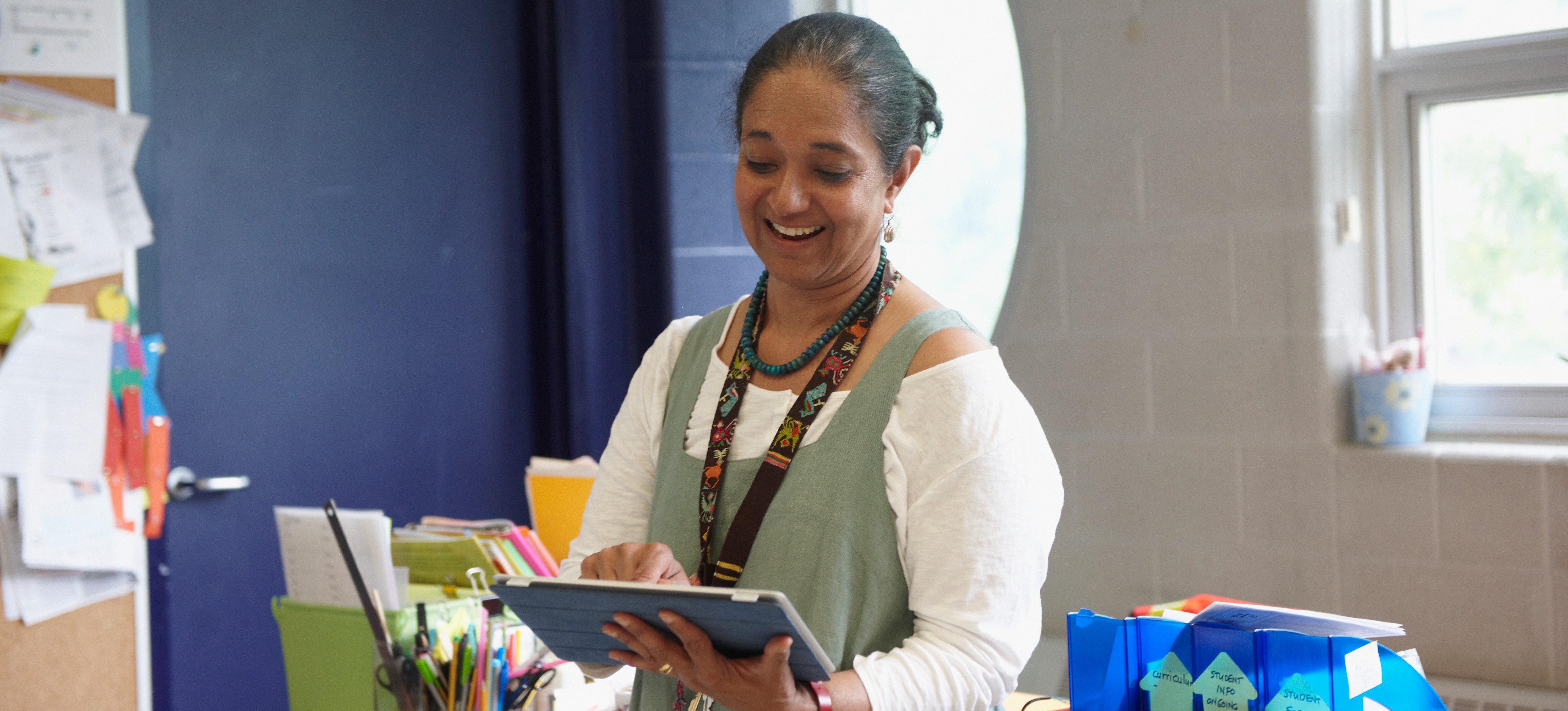 [Featured image] A teacher in a classroom holds a computer tablet as she uses AI for course design ideas.
