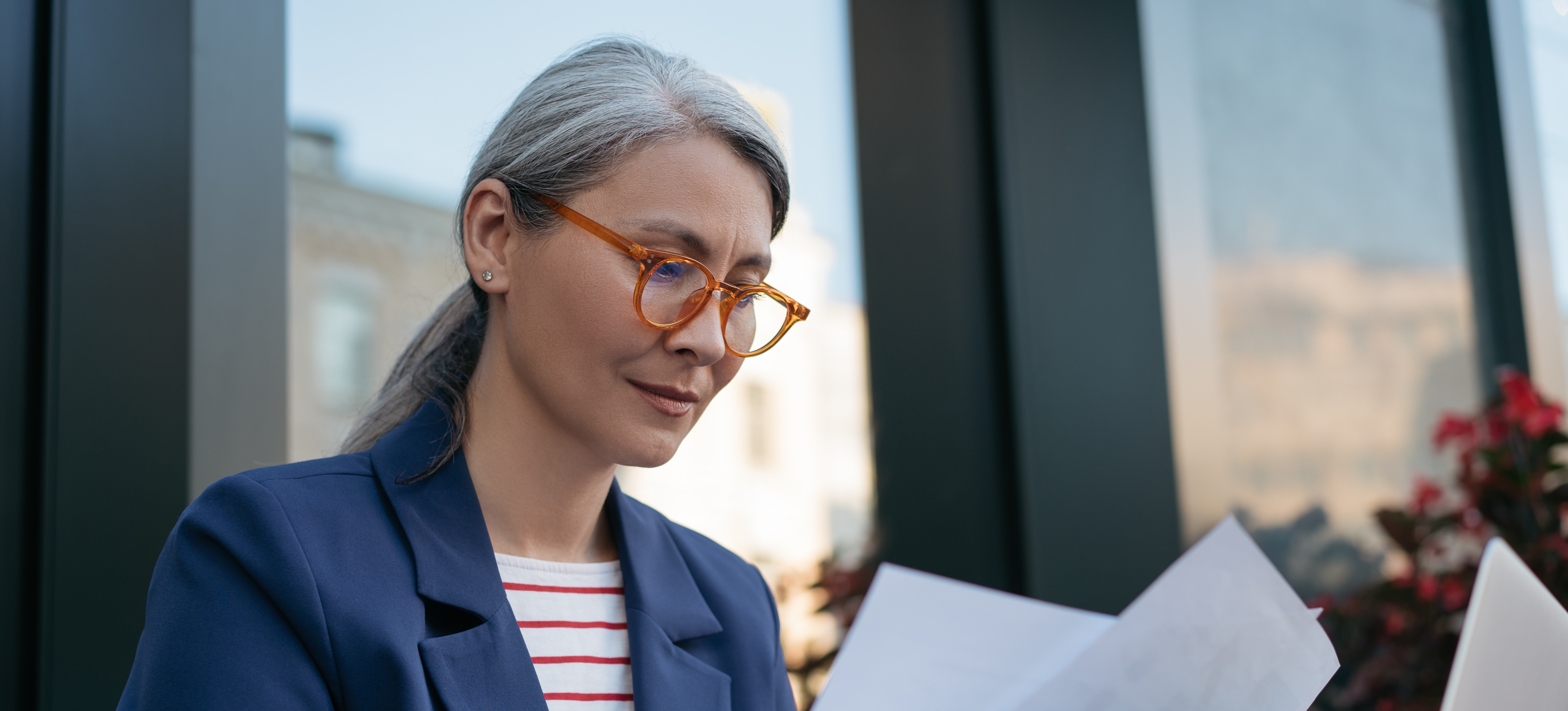 [Featured Image] A person wearing a blue jacket, red and white top, and glasses works on their resume. 
