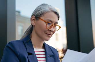 [Featured Image] A person wearing a blue jacket, red and white top, and glasses works on her resume. 