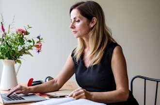 [Featured image] An MBA student attends a lecture on her laptop computer. She's sitting at a table next to a window.