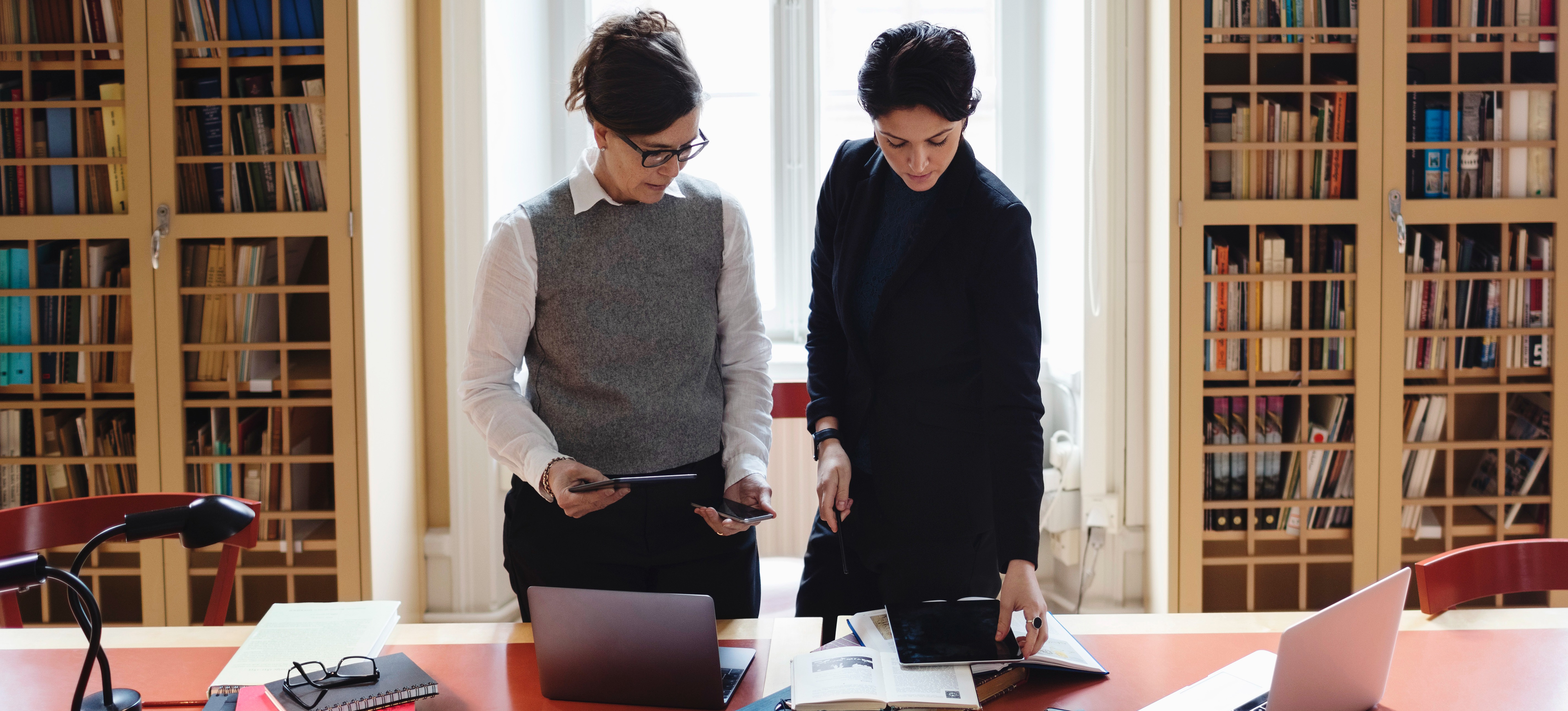 [Featured image] Two accountants who use double-entry accounting look over ledgers on an office table with many bookshelves and a window behind them.