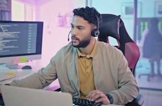 [Featured Image] Man sitting at a desk with several computer monitors in his office, wearing a headset and working intently at his job as a cloud security engineer.