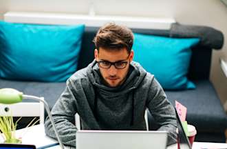 [Featured Image] A person in a dark sweatshirt and glasses sits at a desk and works on a laptop. There is a second laptop on the desk a sofa with two blue pillows behind them. 