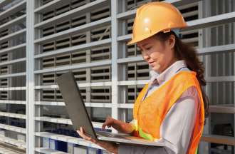 [Featured Image] A woman in an orange vest and hardhat evaluates her company's current supply chain workflow.