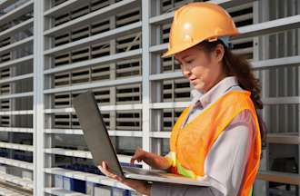 [Featured Image] A woman in an orange vest and hardhat evaluates her company's current supply chain workflow.