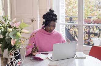 [Featured image] Woman starts her side hustle from home on her laptop while sitting at her dining room table in her light-filled space. She's taking notes in a notebook and sitting beside fresh flowers.