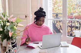 [Featured image] Woman starts her side hustle from home on her laptop while sitting at her dining room table in her light-filled space. She's taking notes in a notebook and sitting beside fresh flowers.