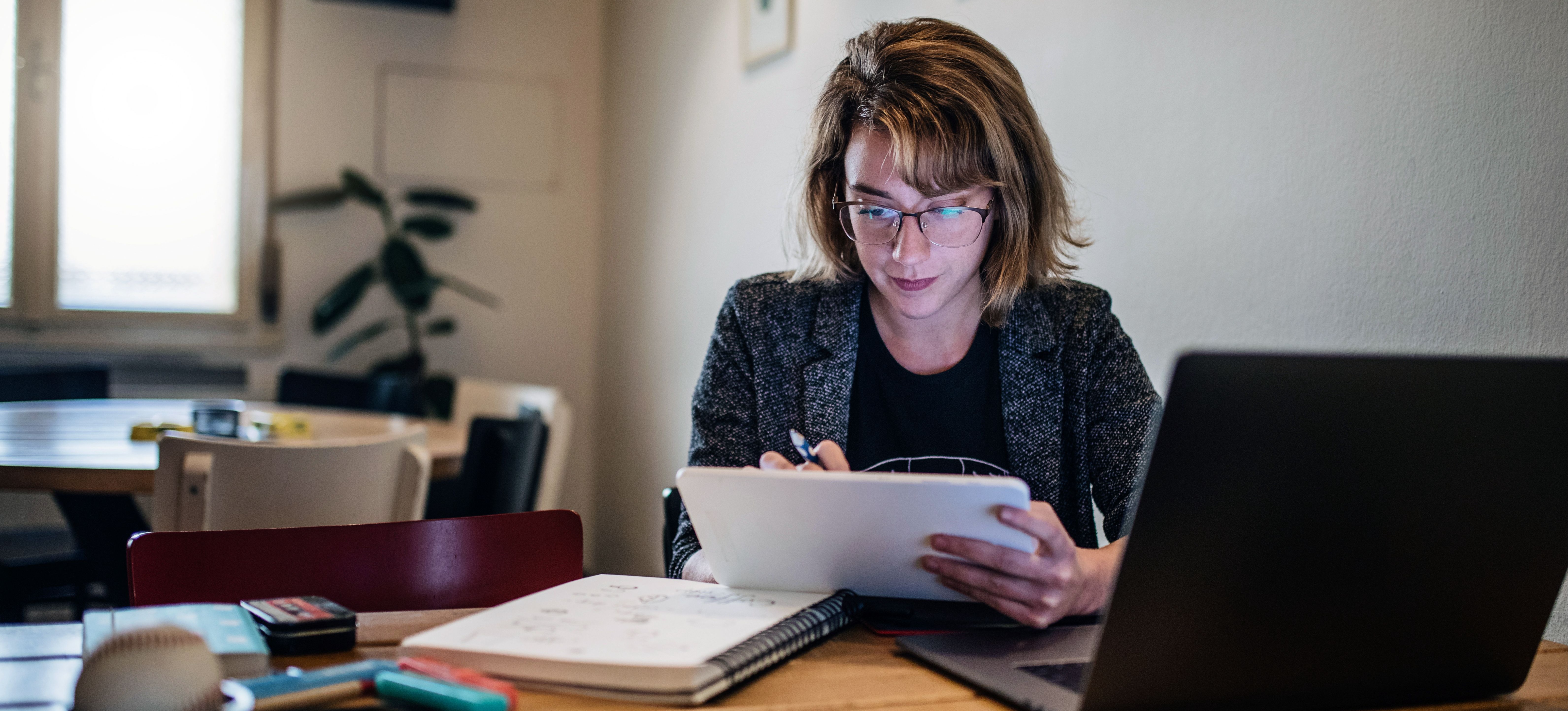 [Featured Image] A person is studiously working on MBA requirements on their tablet and laptop with a notebook and pens on their desk.