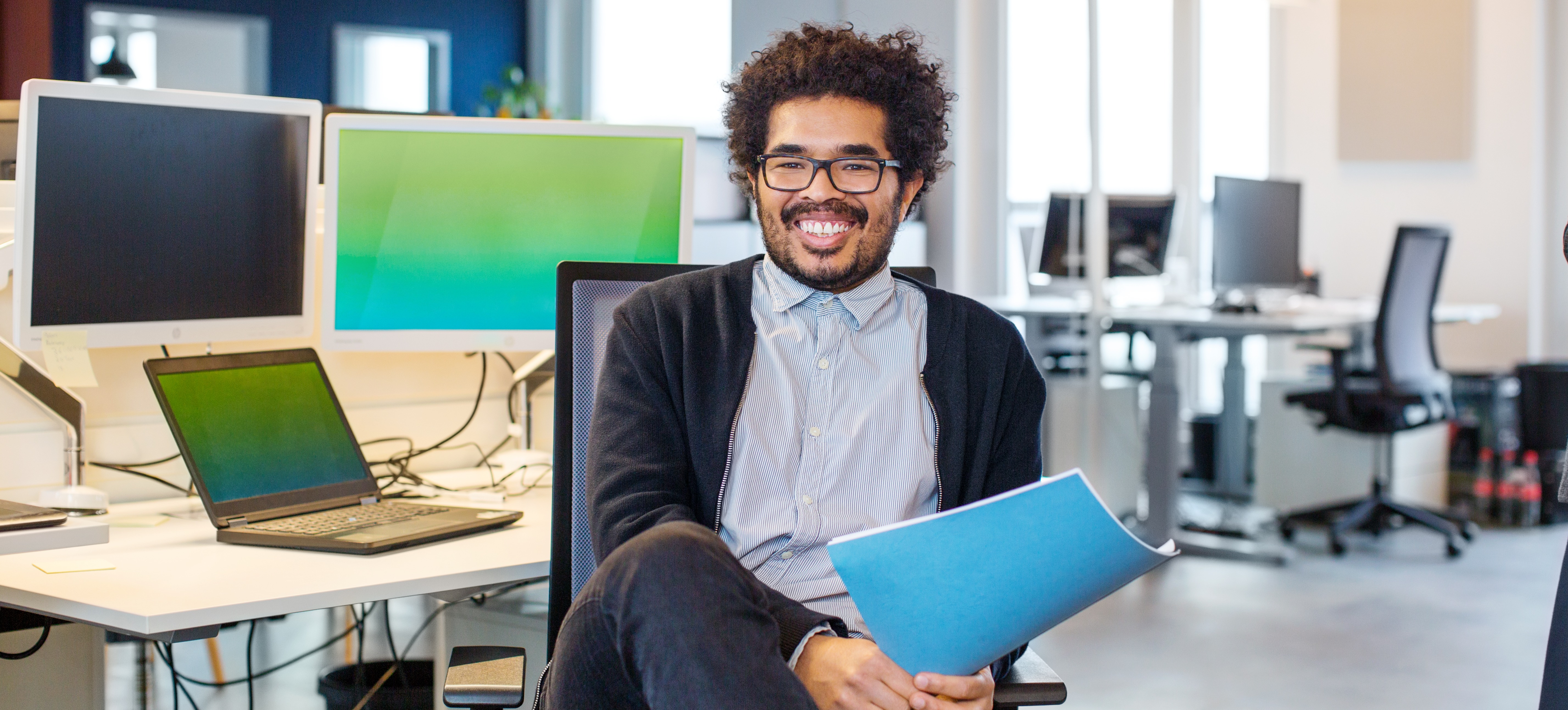 [Hauptbild] Business Analyst hält eine Datei, während er vor einem Laptop und Monitor sitzt