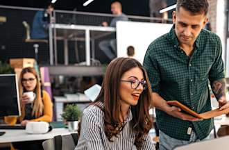 [Featured Image]:   A male, wearing a green shirt and a female wearing a black and white patterned top, sitting in front of her laptop, are meeting to discuss the latest strategy. 