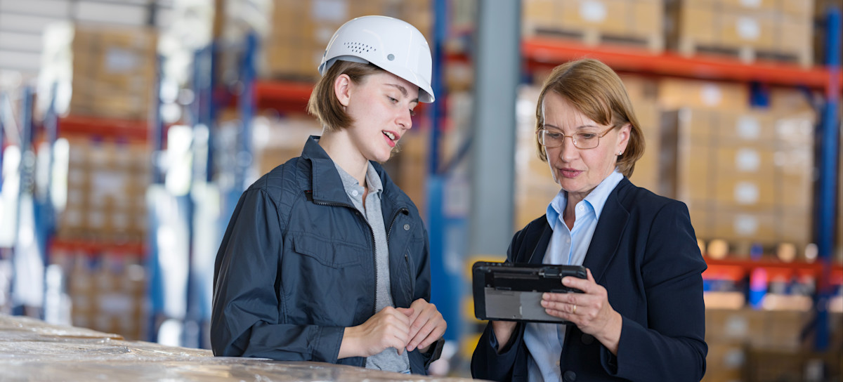 [Featured Image] Two professionals in a warehouse review the potential benefits of implementing AI in supply chain operations. 
