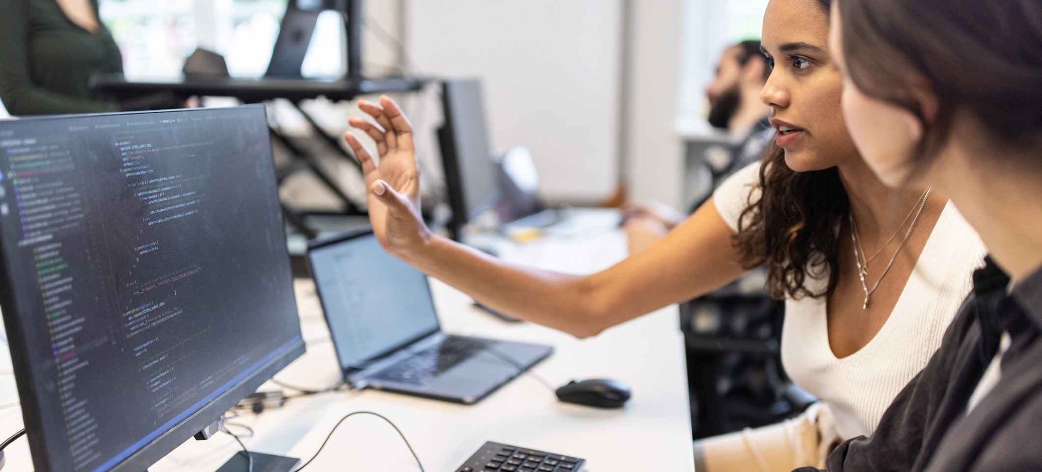 [Featured Image] Professionals in a work environment discussing how to improve Java programming skills while reviewing code on their computers.