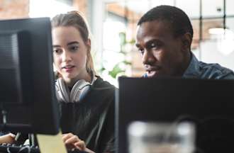 [Featured Image] Two co-workers sit next to each other at a desk and collaborate on a shared computer in an open office space.