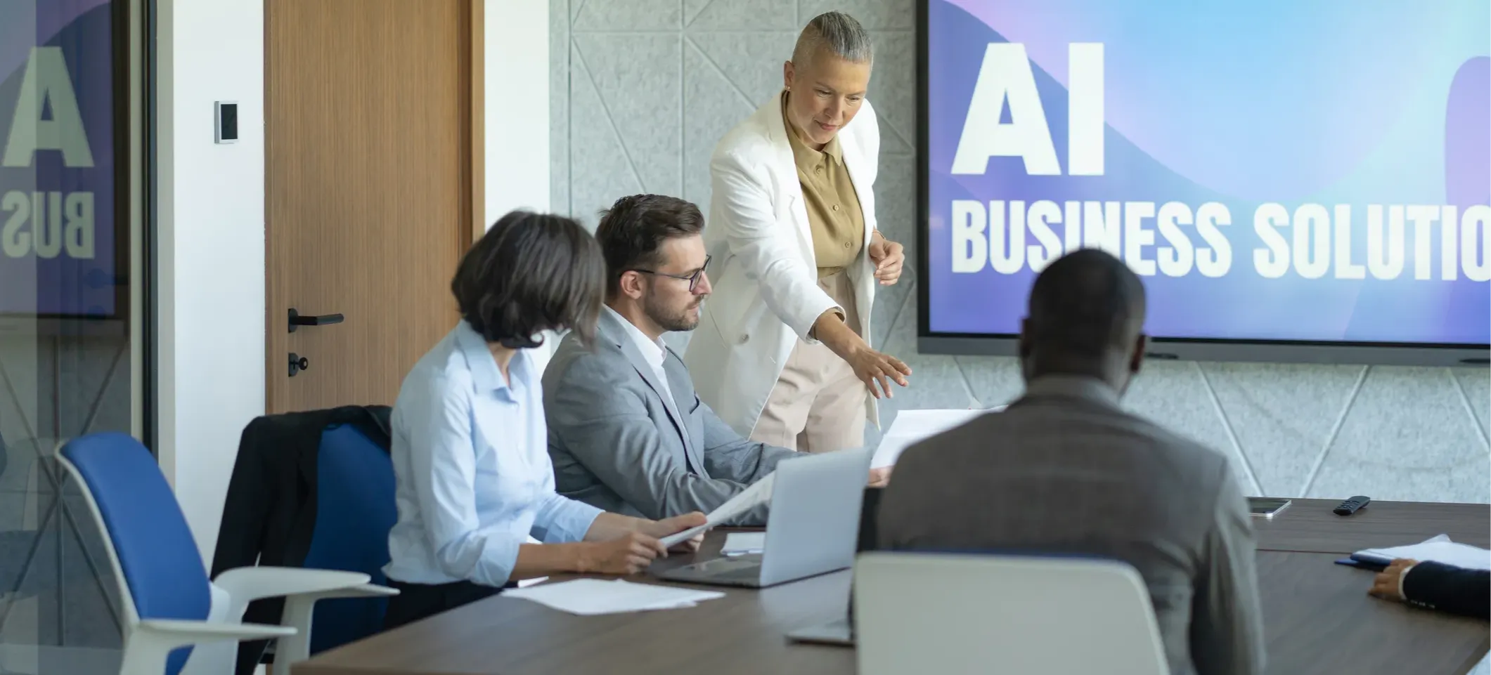 [Featured image] A professional trained in AI strategy consulting stands at the head of a conference room and speaks to a team seated at a table. A large screen is mounted on the wall behind the consultant, displaying the words, "AI business solutions."
