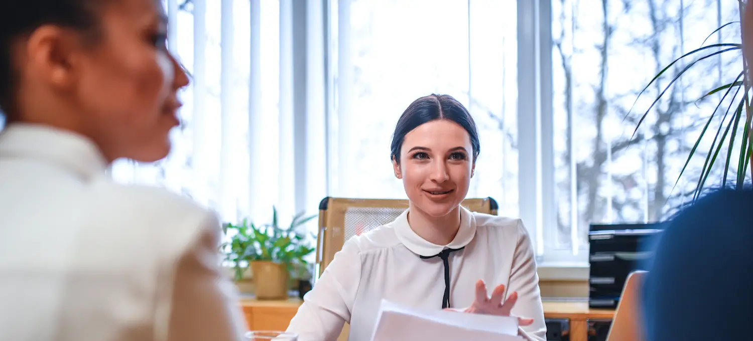 [Featured image]: A financial manager is sitting at their brown desk, holding documents, as they conduct a meeting with two co-workers. 