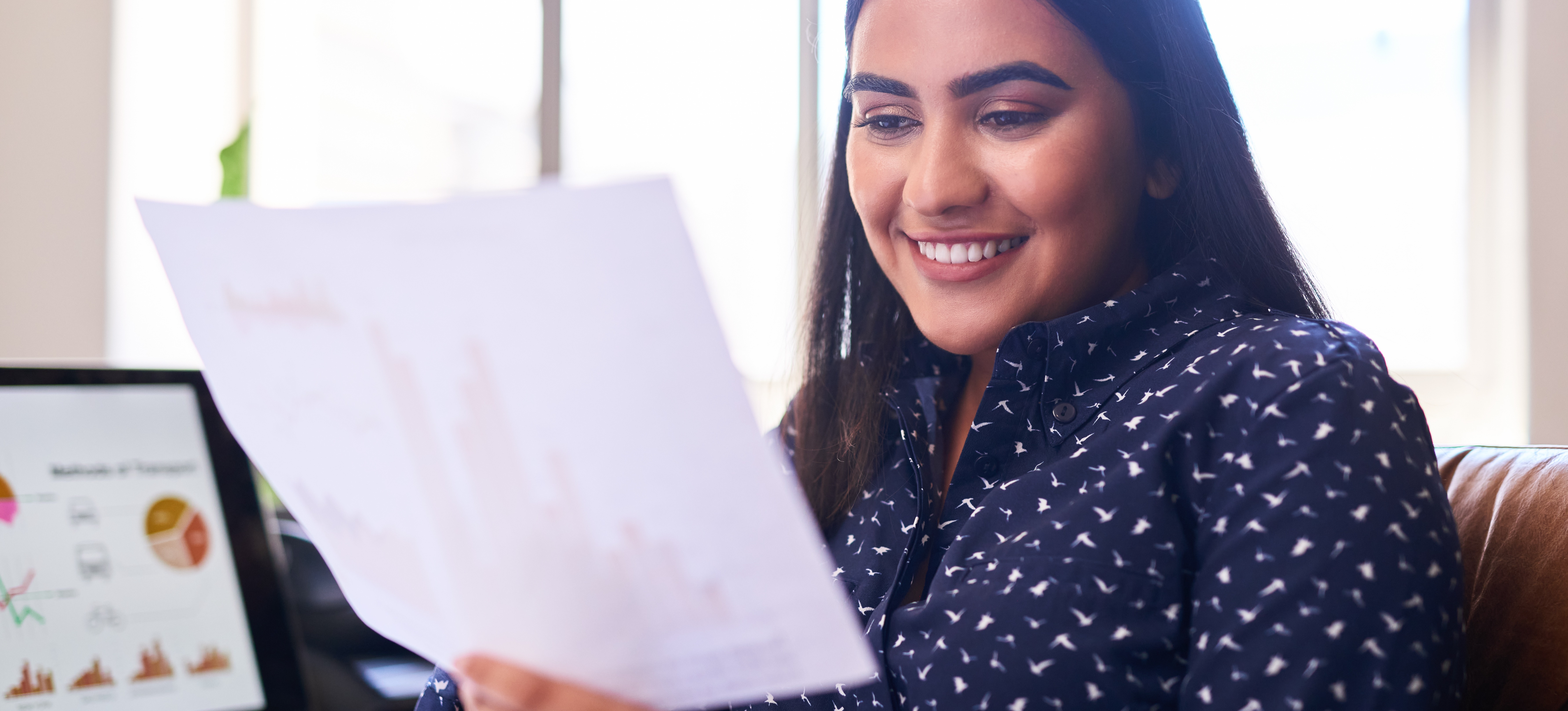 [Featured Image] A smiling digital marketing professional sits at her office desk reading a report with her laptop displaying data in the background.