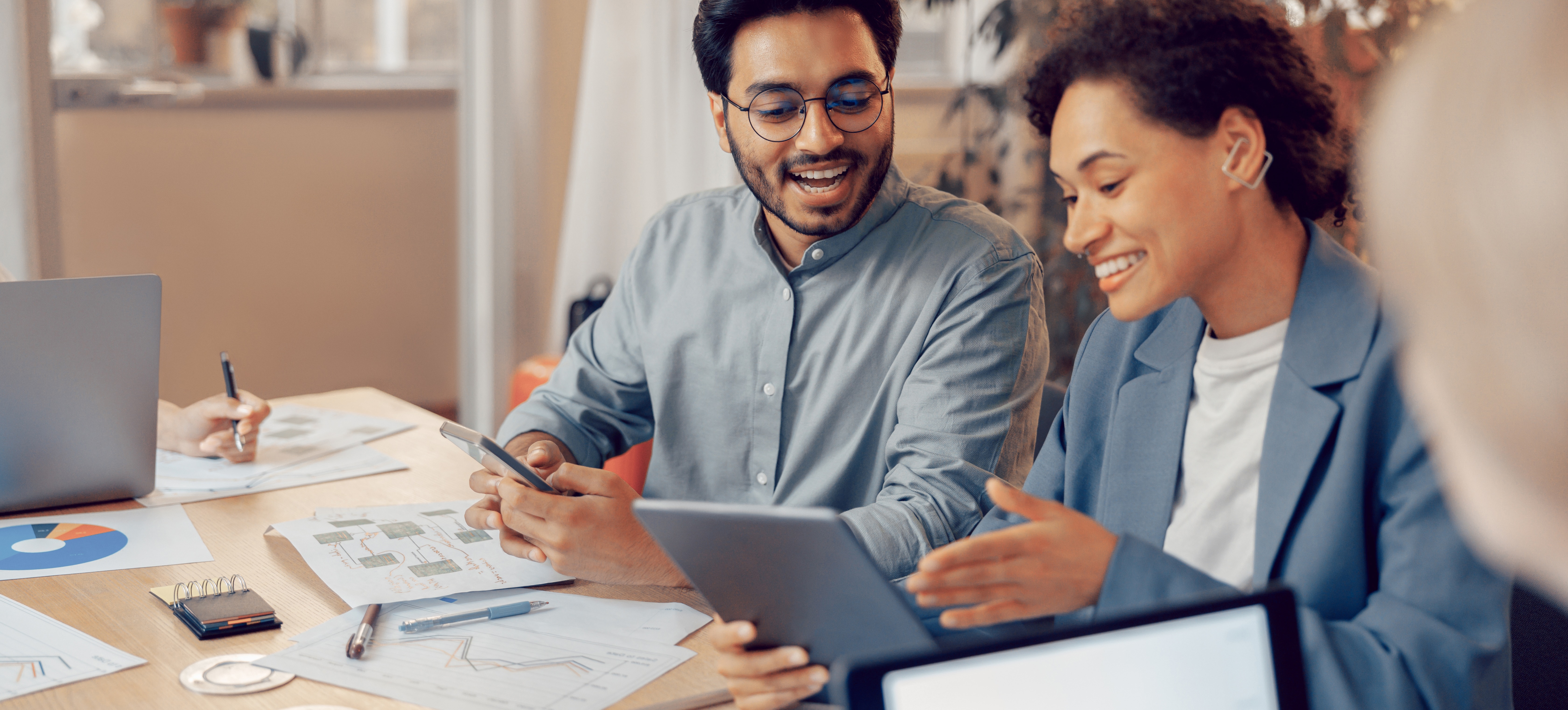 [Featured Image] Four business persons sit at a table in an office, looking at graphs and data and discussing strategies based on business analytics.
